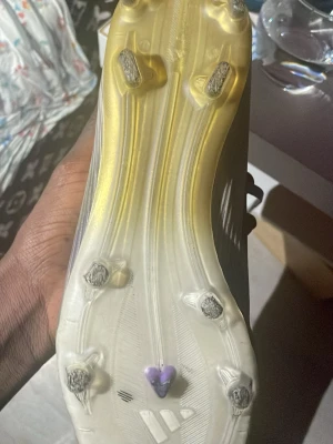 Adidas vita och gula fotbollsskor - Säljer ett par Adidas fotbollsskor med vita och gula detaljer samt lila inslag. Skorna har snörning och en lätt, ribbad ovandel för bra bollkontroll. Sulan har fasta dobbar för spel på naturgräs. Perfekta för dig som vill ha stilrena och snabba skor på planen.