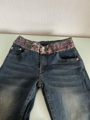 Mörkblå jeans med paljettmidja (nya utan prislapp) - Unika Bootcut jeans med bred midja i paljettmönster i rosa och silver. Bakfickan är dekorerad med glittrande stenar som ger extra bling. Klassisk femficksmodell i stretchigt denim, perfekta för dig som vill sticka ut.