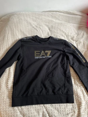 Svart EA7 sweatshirt från Emporio Armani - Snygg svart sweatshirt från EA7 Emporio Armani med guldfärgad logga på bröstet och tryck på ena ärmen. Tröjan har rund halsringning, långa ärmar och ribbade muddar. Perfekt för en trendig och sportig look.