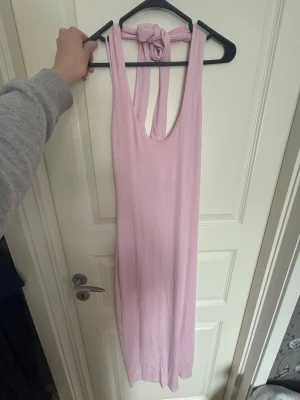 Ljusrosa långklänning med knyt - Säljer en ljusrosa långklänning med djup urringning och breda axelband som knyts i nacken. Klänningen är ärmlös och har en mjuk, följsam passform. Perfekt för varma dagar eller sommarens alla tillfällen. Klänningen är öppen i ryggen