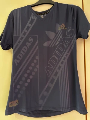 Blå Adidas t-shirt med tryck - Snygg blå t-shirt från Adidas med stort grafiskt tryck och logga framtill. T-shirten har v-ringning och korta ärmar. . Perfekt för dig som gillar sportig och avslappnad stil.