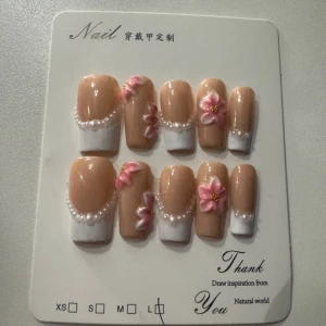 Press-on naglar med pärlor och blommor - Snygga press-on naglar i nude och vitt med 3D-blommor i rosa och små pärlor som dekoration. Naglarna har en elegant, fyrkantig form och passar perfekt för dig som vill ha en söt och trendig look. Materialet är plast.