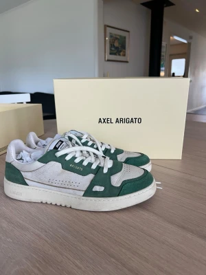 Axel Arigato vita och gröna sneakers - Shyssta sneakers från Axel Arigato, varsamt använda. Säljes på grund av att de har blivit för små. Sand vit och grön färg. Funkar för båda kön. Kartongen ingår, kvitto finns 