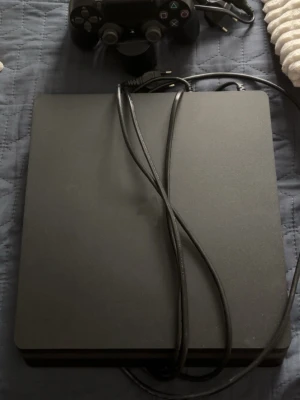 PlayStation 4 Slim med handkontroll - Säljer en svart PlayStation 4 slim från Sony. Konsolen har en stilren, rektangulär design i matt plast och kommer med en matchande svart DualShock 4-handkontroll samt strömkabel. Perfekt för gaming och streaming. Använd i några år men är i fantastiskt skick och funkar alldeles utmärkt. Kommer med 3 spel som finsn på bilden!