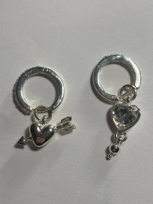 Silverörhängen med hjärtan och pil - Säljer ett par trendiga silvriga örhängen med hjärtformade hängen. Ena örhänget har ett hjärta med pil och det andra har ett hjärta med glittrande sten och små kulor. Båda har rund ring och detaljer i blank metall. Perfekt för dig som gillar unika accessoarer. De går även att ta av från självaste ringen och tex sätta på ett halsband! Andvända få gånger, kommer såklart tvättade