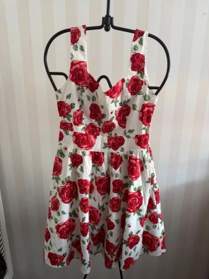 Vit klänning med röda rosor ny  - Söt kortklänning med breda axelband och hjärtformad urringning. Klänningen är vit med stora röda rosor och gröna blad i ett blommigt mönster. Tillverkad i ett lätt bomullstyg som passar perfekt till sommaren. Kjolen är utställd och ger en feminin siluett.