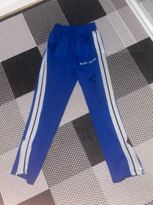 Blåa Palm Angels mjukisbyxor - Snygga blåa mjukisbyxor från Palm Angels med vita och svarta sidoremsor. Byxorna har resår i midjan och dragkedjor vid bensluten. Tillverkade i ett mjukt och stretchigt material som ger en sportig vibe. Perfekta för en avslappnad streetwear-look.