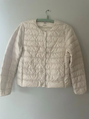 Pufferjacka Uniqlo ultra light down - Lätt och stilren vitbeige pufferjacka från Uniqlo med rund halsringning och tryckknappar framtill. Jackan har quiltad design, långa ärmar och är tillverkad i ett tunt, värmande material som passar perfekt för svensk vår. Finns tre mindre fläckar på insidan av ena ärmen, därav priset. Men inget som direkt syns när jackan är på.
