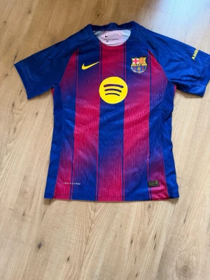 FC Barcelona fotbollströja hemma 25/26 player edition - Säljer en FC Barcelona fotbollströja player edition från nuvarande säsong 25/26 från Nike med klassiska blå och röda vertikala ränder. Tröjan har klubbmärke, Nike-logga och Spotify som sponsor framtill samt UNHCR-logga bak. Tillverkad i lätt och ventilerande material, perfekt för fotbollsträning eller match. Pris kan diskuteras vid snabb affär.