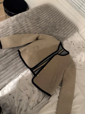 Beige kofta med svarta detaljer - Stilren beige kofta med svarta kanter och guldfärgade knappar. Kortare modell med raka ärmar och två fickor framtill. Perfekt för dig som gillar klassisk men trendig look. Storlek S från Zara aldrig använd.