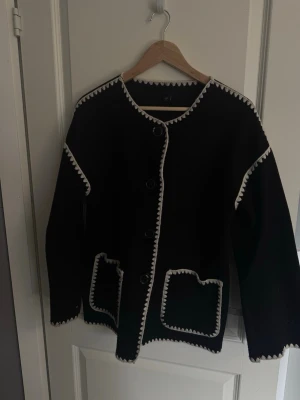 Vårjacka  - Stilren vårjacka kofta med vita virkade kanter längs hals, ärmar och fickor. Jackan har rund halsringning, stora knappar framtill och två framfickor med dekorativa sömmar. 