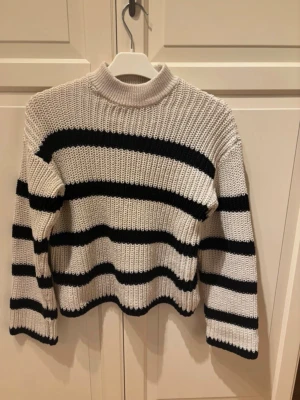 Randig stickad tröja från Vero Moda - Säljer en beige stickad tröja från Vero Moda med svarta horisontella ränder. Tröjan har hög hals, långa ärmar och är ribbstickad för en skön och avslappnad look. Använd max 2 gånger 