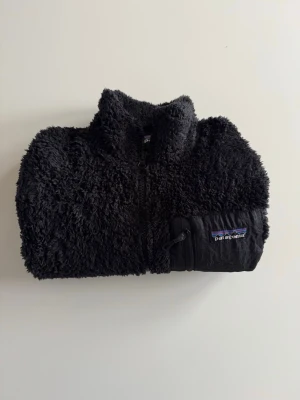 Patagonia, dam, L svart fleecejacka   - Mysig svart teddyjacka från Patagonia med dragkedja framtill och hög krage. Jackan har en bröstficka  med logga och fluffigt material över hela. Perfekt för kalla dagar när du vill vara både varm och stilren.