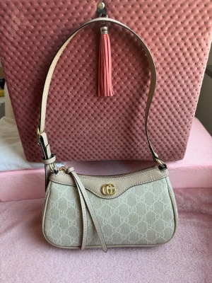 GG Beige axelremsväska - Ny snygg beige axelremsväska från GG. Väskan har guldfärgad logga framtill och dragkedja upptill. 