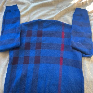 Burberry tröja i kashmire - Säljer en stilren blå tröja från Burberry med ett klassiskt rutmönster i rött och svart. Tröjan är långärmad och tillverkad i Kashmir och bomull, vilket gör den både bekväm och elegant. Perfekt för att ge en touch av lyx till din garderob. Skriv om ni har några frågor