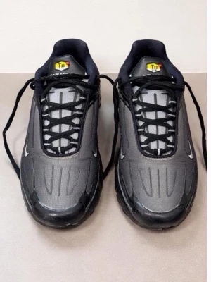 Nike Tuned Air svarta sneakers - Nike Tuned Air sneakers i svart och mörkgrått med vita detaljer. Skorna har en sportig design med synliga sömmar, svarta snören och Tn-logga på plösen. Ovandelen är i syntetmaterial och mesh för bra andningsförmåga. Perfekta för dig som gillar streetwear.