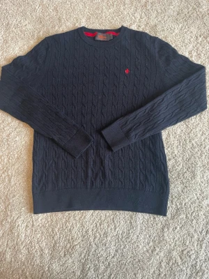 Mörkblå kabelstickad tröja Morris  - Snygg mörkblå kabelstickad tröja från Morris med rund hals och långa ärmar. Tröjan har ett rött broderat emblem på bröstet och ribbade muddar vid ärmslut och nederkant. Perfekt för dig som gillar klassisk och stilren design.