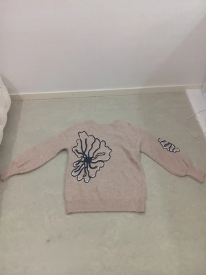 Beige stickad tröja med brodyr - Beige stickad tröja med unika broderade blommor i svart och på ryggen och ärmarna. Tröjan har rund halsringning, ribbade muddar och en avslappnad passform. Perfekt för dig som vill ha något personligt och trendigt i garderoben.