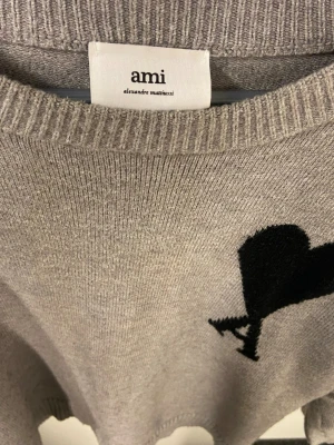 Grå stickad tröja från Ami Paris - Snygg grå stickad tröja från Ami Paris med svart hjärta och bokstaven A broderat på bröstet. Tröjan har rund halsringning, ribbade muddar och är tillverkad i mjukt material som passar perfekt till en avslappnad stil. Passar i både kön!!! Jag rekommenderar att du köpet det snabbt innan en annan gör! 