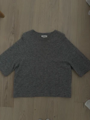 Grå stickad tröja från & Other Stories - Mysig grå stickad tröja från & Other Stories med rund halsringning och halvlånga ärmar. Tröjan har en enkel, rak passform och är tillverkad i ett mjukt material som passar perfekt för kyligare dagar.