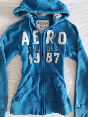 Blå zip hoodie från Aeropostale - Passar mig som har xs. Blå hoodie med dragkedja från Aeropostale. Stor vit brodyrtext 'AERO' och siffrorna 1987 framtill. Har två fickor, huva med dragsko och mjukt innerfoder i grått. 