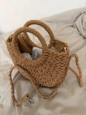 Flätad beige handväska med axelrem - Säljer en superfin handväska i flätad strådesign med rundade handtag och avtagbar axelrem. Väskan är beige och har en mjuk, naturlig look som passar perfekt till sommaren. Insidan är fodrad och väskan stängs med dragkedja. Perfekt accessoar för en trendig outfit.