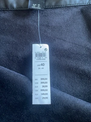 Ny Svart volangkjol i skinnimitation - Snygg svart långkjol i skinnimitation med volangdetalj nedtill. Kjolen har en hög midja och en stilren, modern look som passar perfekt till olika outfits. Materialet ger en cool känsla och volangen gör kjolen extra trendig.