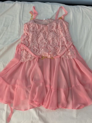 Rosa spetsklänning från Ida Sjöstedt - Supersöt rosa klänning från Ida Sjöstedt med spetsdetaljer upptill och volangig kjol i chiffong. Klänningen har smala axelband och dekorativa guldfärgade detaljer. Perfekt för sommarens alla tillfällen!