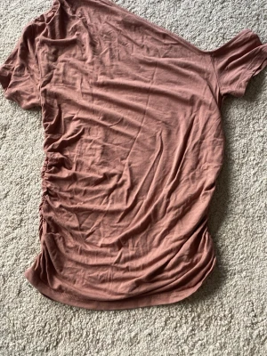 Brun oneshoulder topp från Gina Tricot - Säljer en brun oneshoulder topp från Gina Tricot i mjuk bomull. Toppen har en asymmetrisk design med rynkade detaljer på sidan och är perfekt för dig som gillar trendiga snitt och enkel stil.