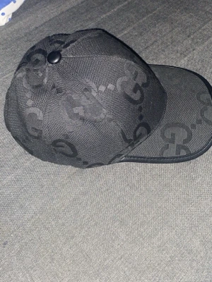 Svart keps från Gucci med GG-mönster - Säljer en svart keps  med det klassiska GG-monogrammet i ton-i-ton över hela kepsen. Kepsen har böjd skärm och justerbar baksida. Materialet är slitstarkt tyg som ger en exklusiv känsla och passar perfekt till streetwear-stilen