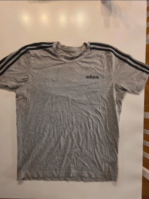Grå Adidas t-shirt med svarta ränder - Snygg grå t-shirt från Adidas med klassiska svarta ränder längs axlarna och svart Adidas-logga på bröstet. T-shirten har rund halsringning och är kortärmad. Perfekt för en sportig och avslappnad stil. STORLEK M  Bröstmått: ca 33cm Längdmått: ca 66 cm