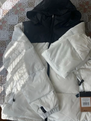 Vit och svart pufferjacka med huva - Snygg pufferjacka i vitt med svart axelparti och huva. Jackan har dragkedja framtill, två sidofickor med dragkedja och elastiska muddar vid ärmsluten. Materialet är glansigt och quiltat för extra värme. Perfekt för kalla vinterdagar.