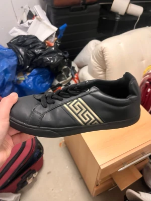 sneakers  - Snygga svarta sneakers från Pier One med guldfärgade geometriska detaljer på sidan. Skorna har klassisk låg profil, svarta skosnören och är tillverkade i slätt konstläder. Perfekta för dig som gillar stilrena och trendiga skor.