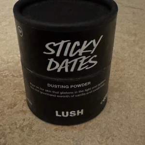 LUSH Sticky Dates Dusting Powder - LUSH Sticky Dates Dusting Powder i svart rund burk. Pudret ger huden en glittrande finish och har en varm doft av vanilj och kakao. Perfekt för att fräscha upp huden och ge en mjuk känsla. Snygg och stilren förpackning med vit text. Användt 2 gånger