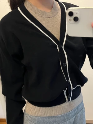 Svart kofta med vita kanter från Mango - Stilren svart kofta från Mango med vita kontrastkanter längs knäppningen, ärmsluten och nederkanten. Koftan har v-ringning, långa ärmar och stängs med fyra knappar framtill. Perfekt att slänga över en t-shirt för en clean look.