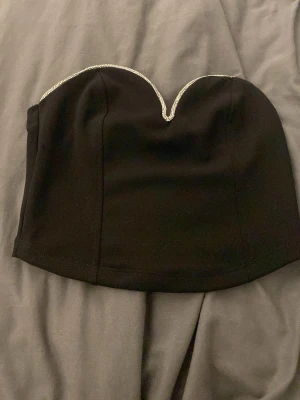 Svart axelbandslös tubtopp med strass - Snygg svart axelbandslös tubtopp med hjärtformad urringning och glittrande strassdetaljer längs kanten. Perfekt att styla till höga jeans eller kjol för en trendig look.