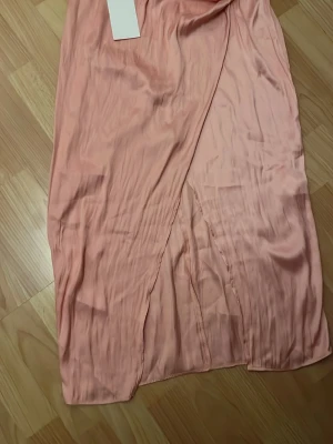 Lång rosa omlottklänning i satin - Säljer en lång rosa omlottklänning i mjukt satinliknande material. Klänningen har en elegant slits framtill och en fin glans i tyget. Perfekt för dig som vill ha en feminin och trendig look.
