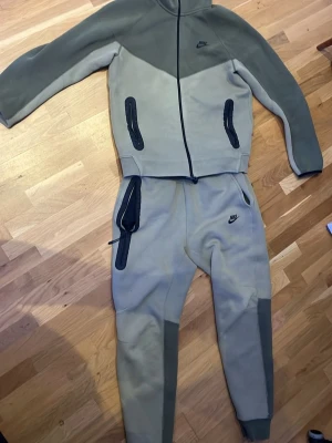 Nike Tech - Säljer ett snyggt loungewear set från Nike i två nyanser av grått. Setet består av en zip-hoodie med svarta dragkedjor och matchande byxor med fickor och Nike-logga. Tillverkat i mjukt material, perfekt för chill dagar hemma.