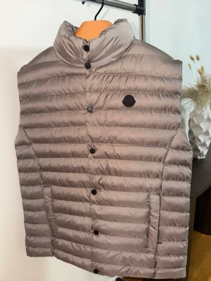 Beige dunväst från Moncler - Snygg beige dunväst från Moncler med diskret svart logotyp på bröstet. Västen har horisontella sömmar, hög krage och knäppning framtill med svarta knappar. Två fickor med dragkedja på sidorna. Perfekt för lager-på-lager och streetstyle.