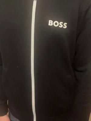 Svart zip-hoodie från BOSS - Svart hoodie med dragkedja från BOSS. Klassisk design med vit logga på bröstet och vit dragkedja framtill. Perfekt för en stilren och avslappnad look. Mjukt material och bekväm passform.