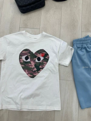 Vit Comme des Garçons t-shirt - Vit t-shirt från Comme des Garçons med stort hjärta i camouflage-mönster och ögon på bröstet. Liten röd hjärtdetalj ovanpå. Klassisk passform och rund hals. Perfekt för dig som gillar streetwear och vill sticka ut.