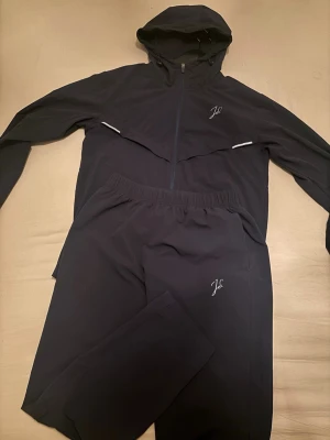 Svart träningsset med huva från jeu d'ombres - Säljer ett blått träningsset från Jeu d’ombres med matchande jacka och byxor. Jackan har huva, hel dragkedja och diskret logga på bröstet. Byxorna har resår i midjan och logga på benet. Perfekt för dig som gillar sportig och stilren look även reflex på loggorna.