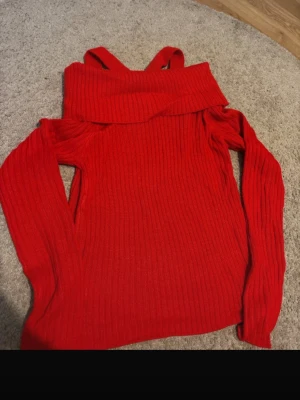 Röd off shoulder stickad tröja - Trendig röd stickad tröja med off shoulder-design och bred krage. Tröjan har långa ärmar och ribbad struktur, samt dekorativa axelband som ger en unik look. Perfekt för dig som vill sticka ut lite extra. Materialet känns mjukt och stretchigt.