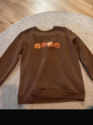 Brun sweatshirt med pumpor - Cool brun sweatshirt med fyra Halloween-pumpor tryckta på bröstet. Tröjan har rund halsringning och långa ärmar. Perfekt för dig som vill ha en höstig vibe och gillar Halloween-tema. Materialet känns mjukt och bekvämt mot huden.