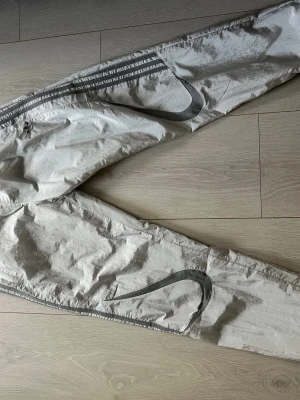 adidas Originals Adicolor 2000s Woven träningsbyxor - Snygga silvriga träningsbyxor från adidas med stora reflex-Swoosh längs benen. Byxorna har resår i midjan och dragkedja vid fickan. Perfekta för en sportig och avslappnad stil. Materialet är mjukt och bekvämt, passar till chill eller träning.