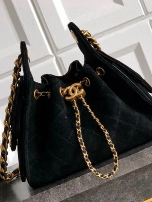 Svart quiltad bucket bag med kedja - Snygg svart bucket bag i quiltat tyg med guldfärgade kedjedetaljer och dragskostängning. Väskan har en axelrem i kombinerat läder och kedja, samt guldfärgade metalldetaljer. Perfekt för dig som gillar trendiga accessoarer.