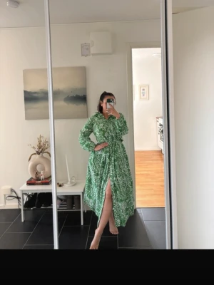 Grön långklänning med bladmönster - Säljer en grön långklänning med vitt bladmönster. Klänningen har lång ärm, v-ringning och markerad midja med knytband. Den har en slits framtill och volangdetaljer upptill. Perfekt för dig som vill ha en färgstark och trendig look.