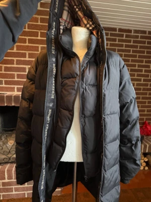 Svart pufferjacka från Burberry - Säljer en svart pufferjacka från Burberry med klassiskt rutigt foder i huvan och logotypband längs dragkedjan. Jackan har en oversized look, dragkedja framtill och stora vadderade sektioner som håller dig varm. Perfekt för kalla vinterdagar.