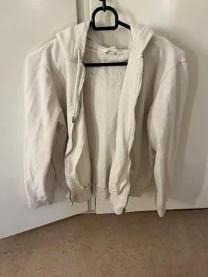 Vit hoodie med dragkedja - Klassisk vit hoodie med dragkedja framtill och huva. Tillverkad i mjukt bomullsmaterial med ribbade muddar vid ärmslut och nederkant. Perfekt för en avslappnad och enkel stil.