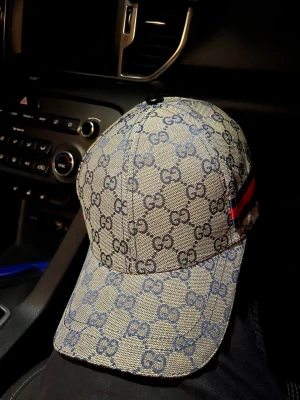 Gucci keps med GG-mönster - Säljer en snygg keps från Gucci med klassiskt GG-monogram i blått på beige botten. Kepsen har böjd skärm och en ikonisk röd-blå rand på sidan. Tillverkad i canvasmaterial som ger en exklusiv känsla och streetwear-vibe.
